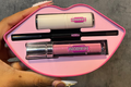 Pink lip-shaped case with makeup items including a lip gloss and eyeliner, held by a hand against a gray background.