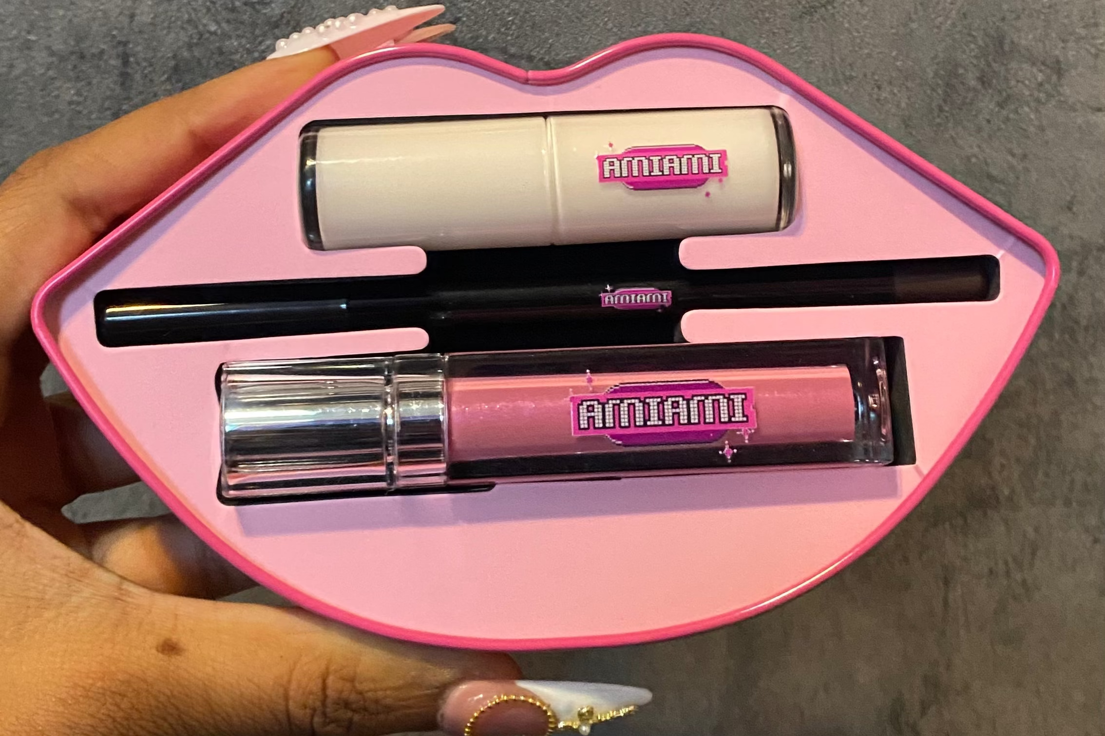 Pink lip-shaped case with makeup items including a lip gloss and eyeliner, held by a hand against a gray background.